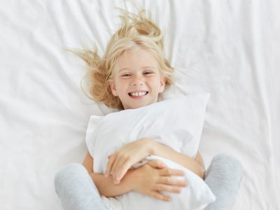 Smiling blonde girl embracing white pillow while being in kindergarten, having good mood while seein
